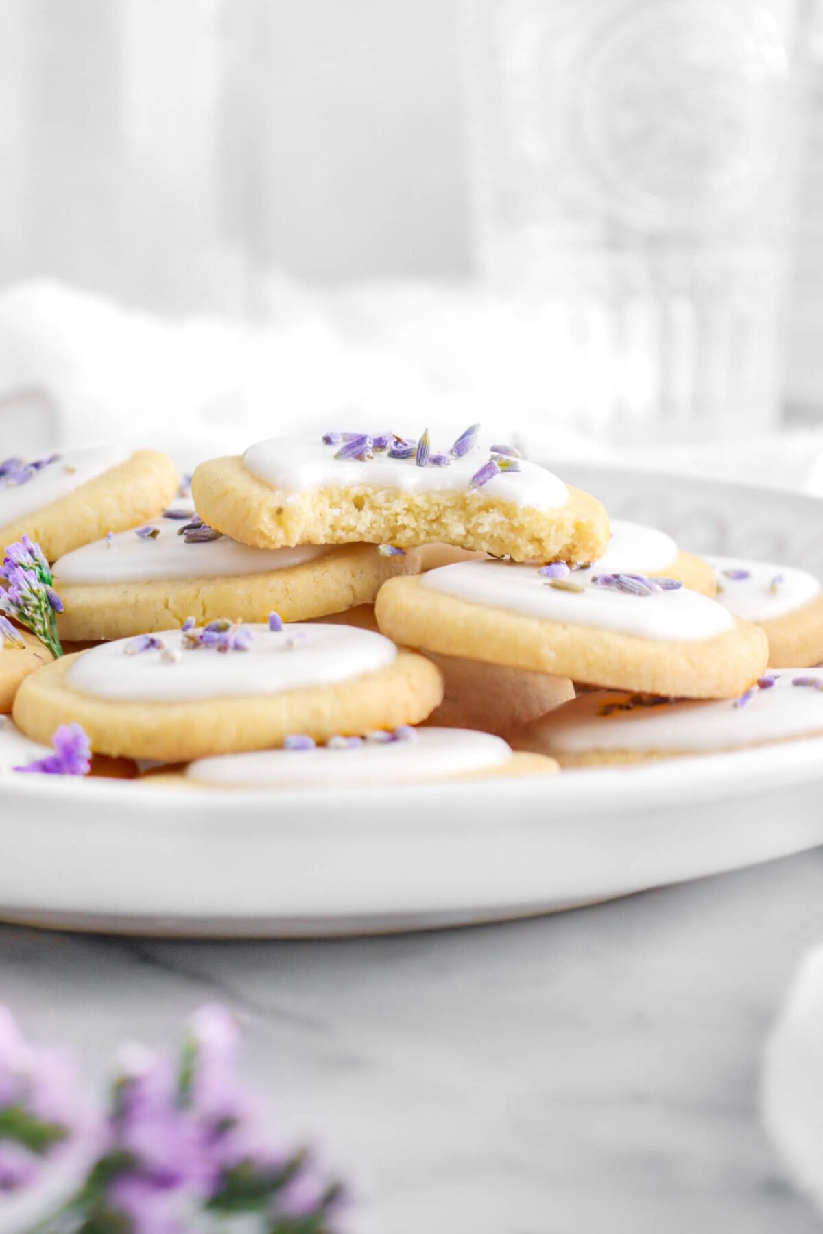 close up front shot of piled lavender cookies on white plate with one cookie missing a bite.