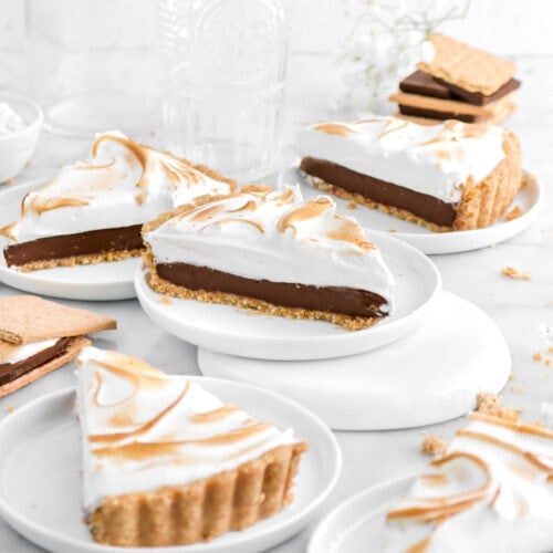 five slices of s'mores pie on small white plates with white flowers and stack of graham crackers and chocolate squares behind, with two empty glasses.