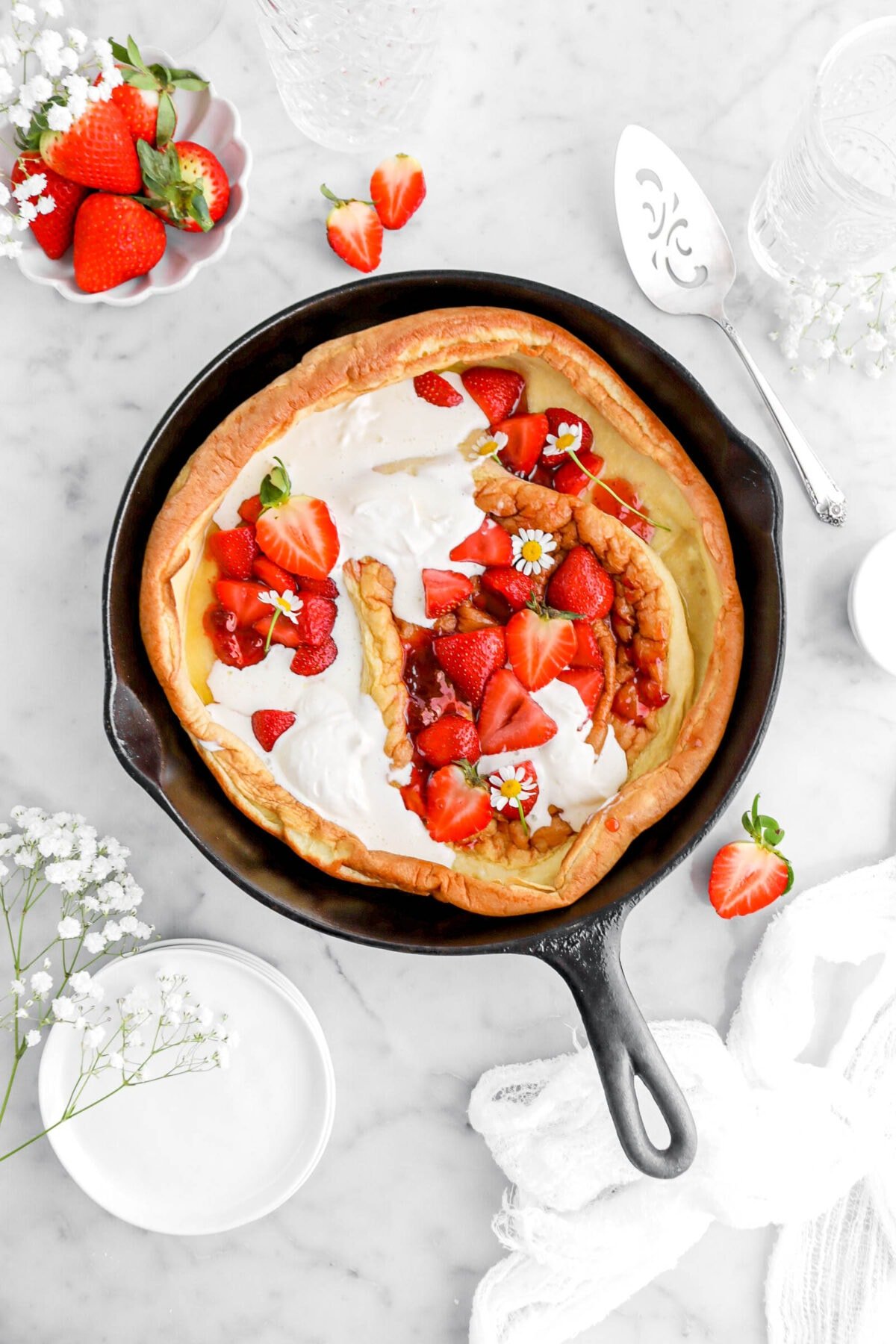 Sautéed strawberries and whipped cream in dutch baby with fresh strawberries and flowers around on marble surface.