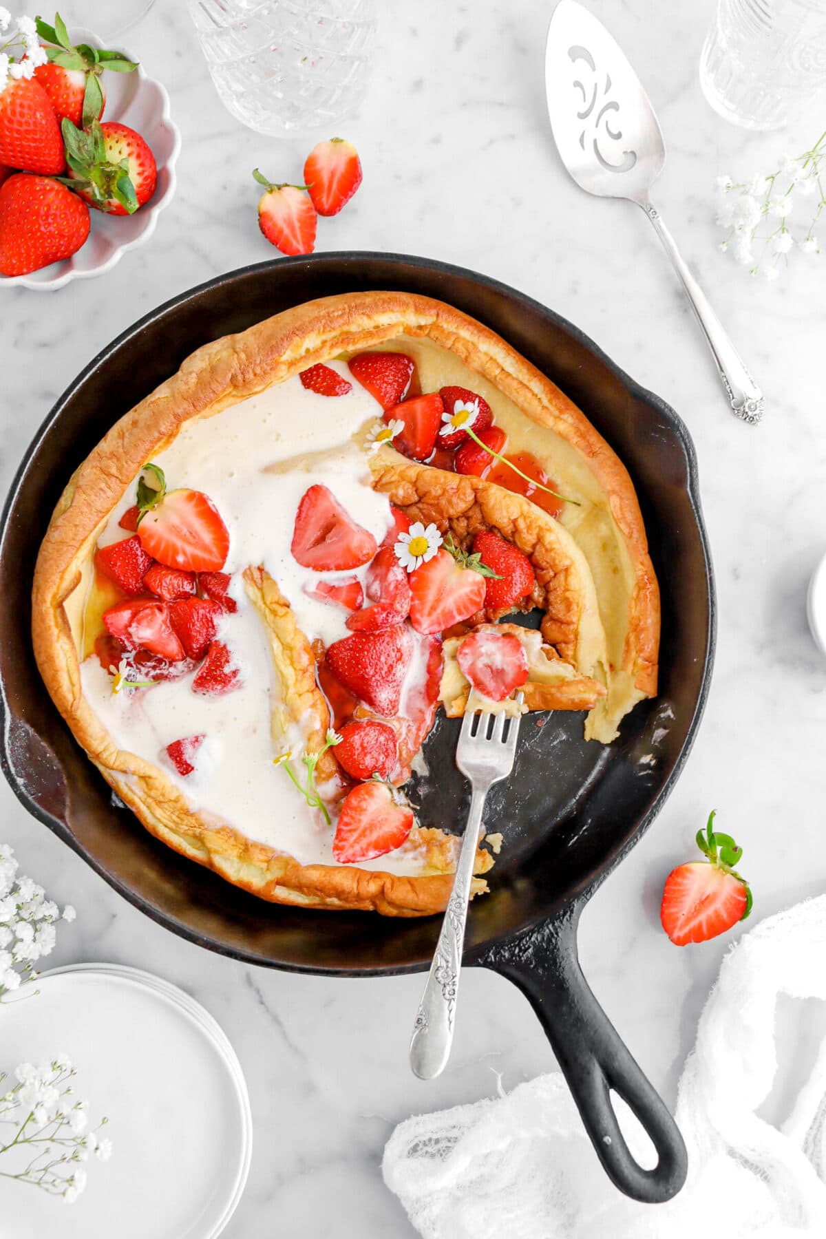 cropped close up of strawberries and cream dutch baby with piece missing and fork stuck into pancake, with fresh strawberries and white flowers around on marble surface.