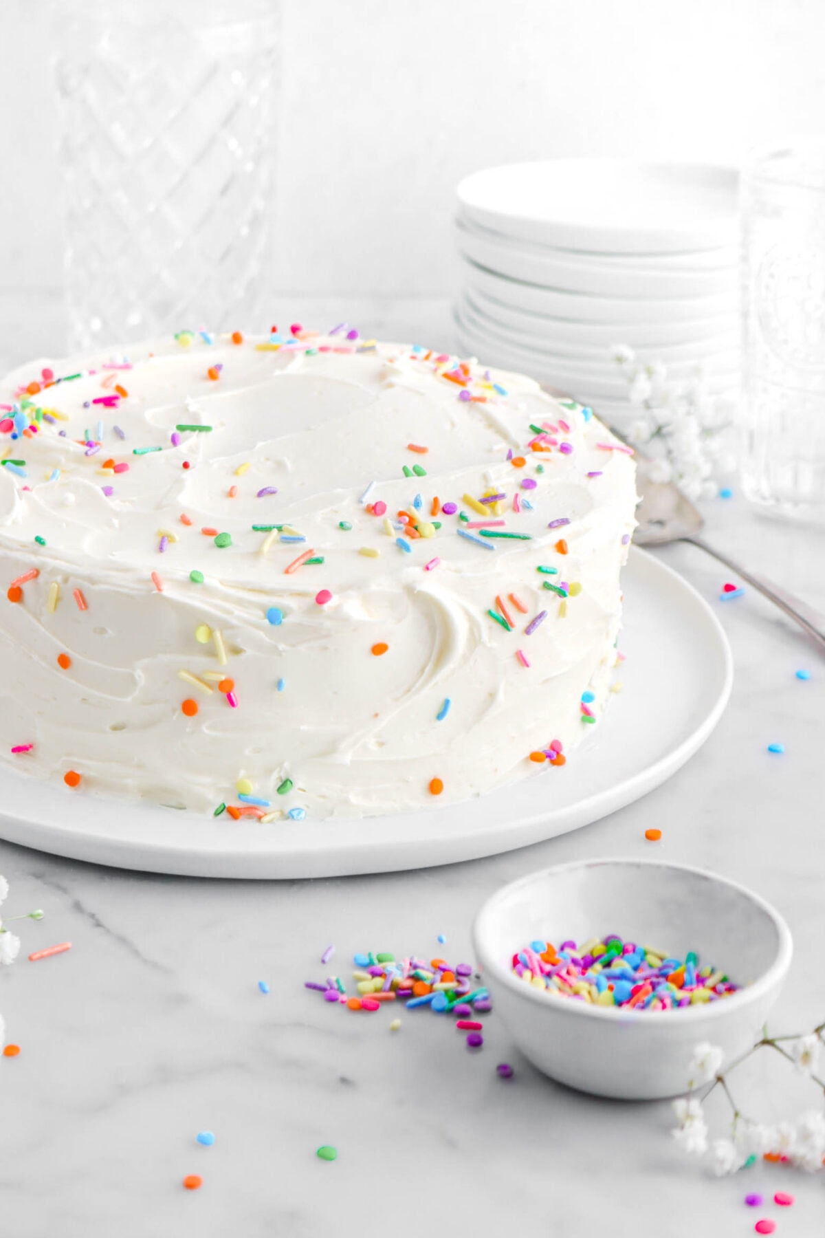 cropped close up of frosted cake with sprinkles on top and sides of cake, with bowl of sprinkles and white flowers around on marble surface.