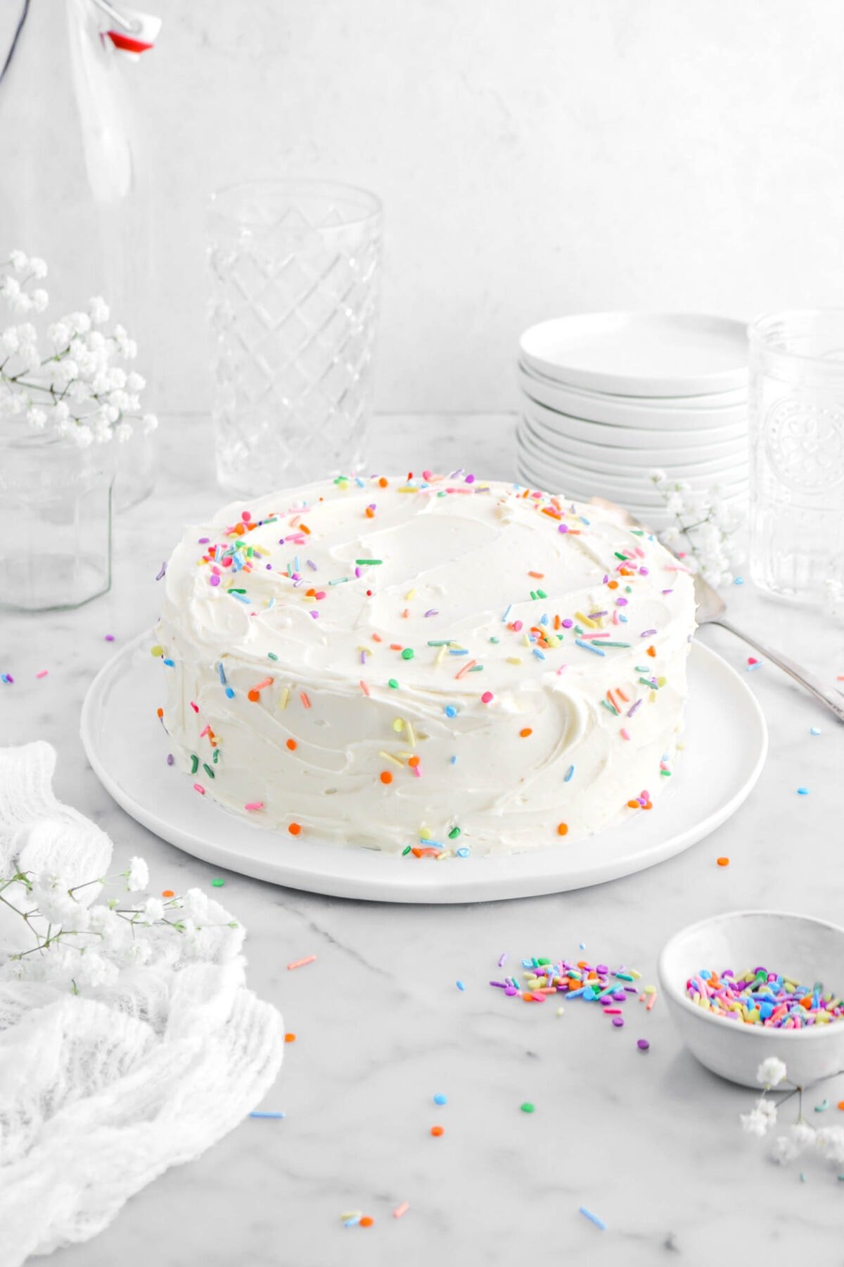front shot of frosted cake with sprinkles scattered around on marble surface, with white cheesecloth beside, white flowers, empty glasses, and stack of plates behind on marble surface.