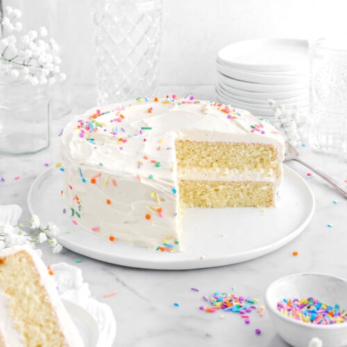frosted vanilla cake on white plate with slice in front ons mall white plate on top of a white cheesecloth with sprinkles and white flowers around.