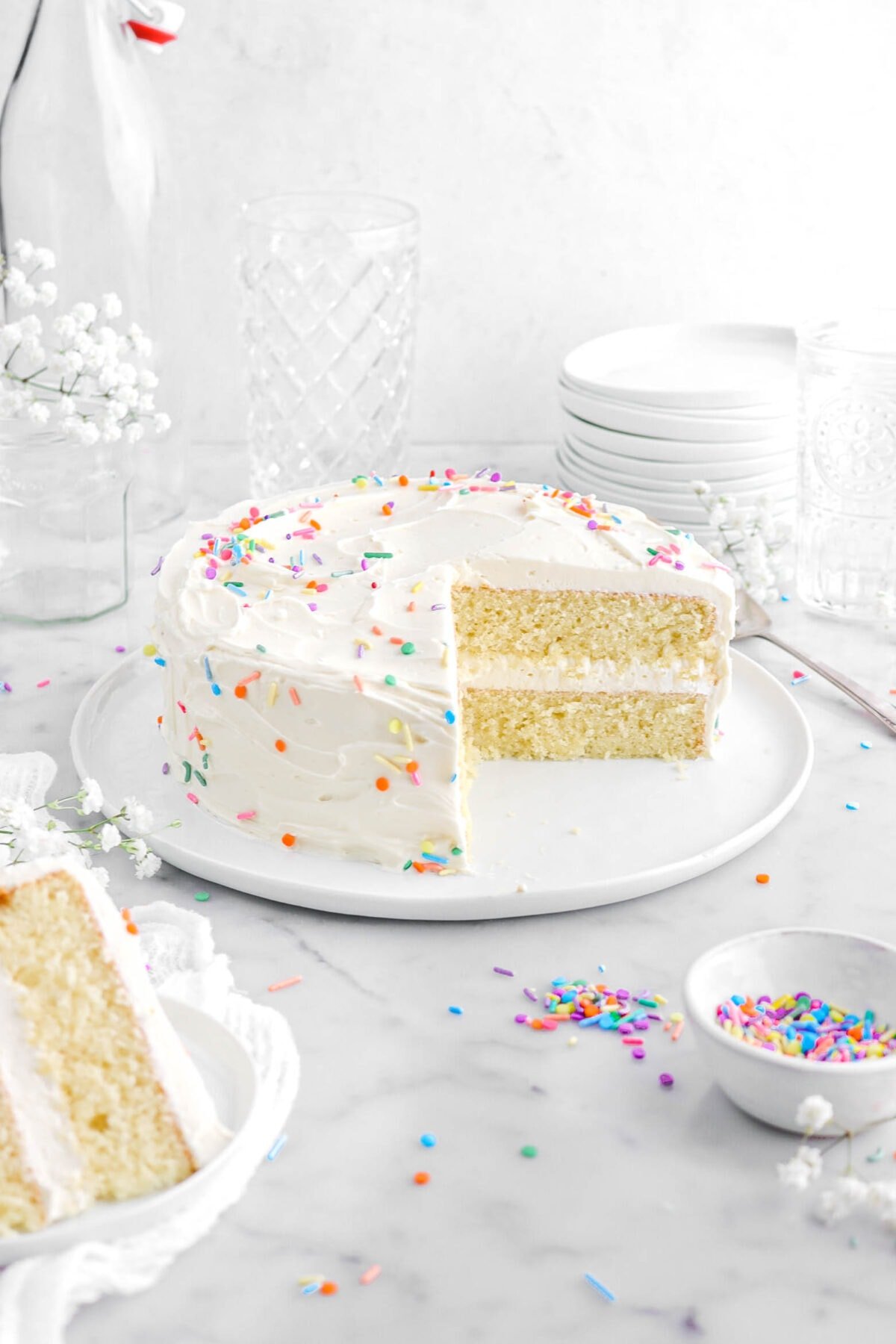 frosted vanilla cake on white plate with slice in front ons mall white plate on top of a white cheesecloth with sprinkles and white flowers around.