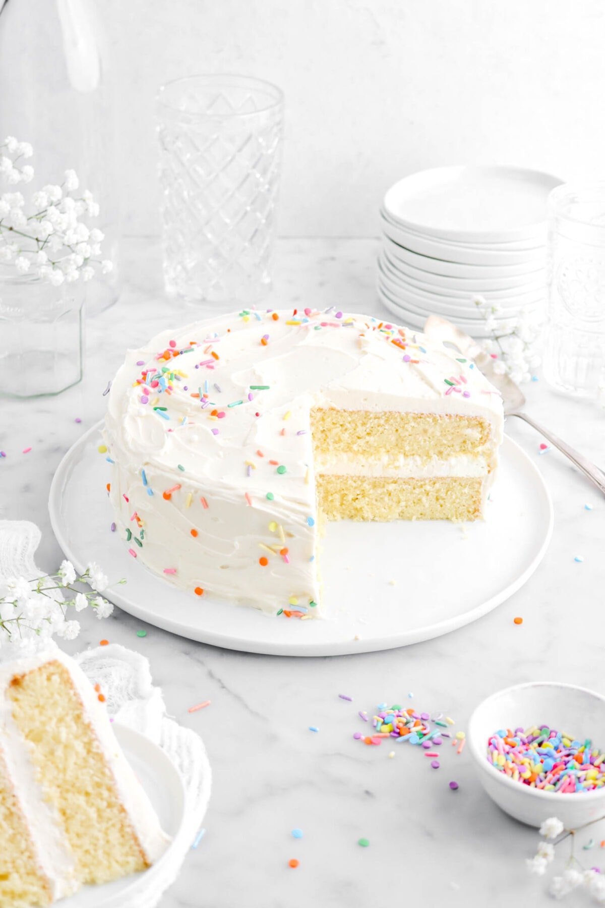 angled shot of cut vanilla cake on white plate with a slice of cake on white plate in front of cake with sprinkles and white flowers around.