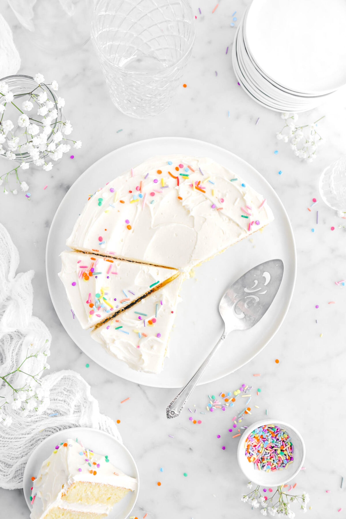 overhead shot of vanilla cake on white plate with two slices cut into and cake knife beside cake, with sprinkles, white flowers, stack of plates, and slice of cake on small white plate on top of white cheesecloth.