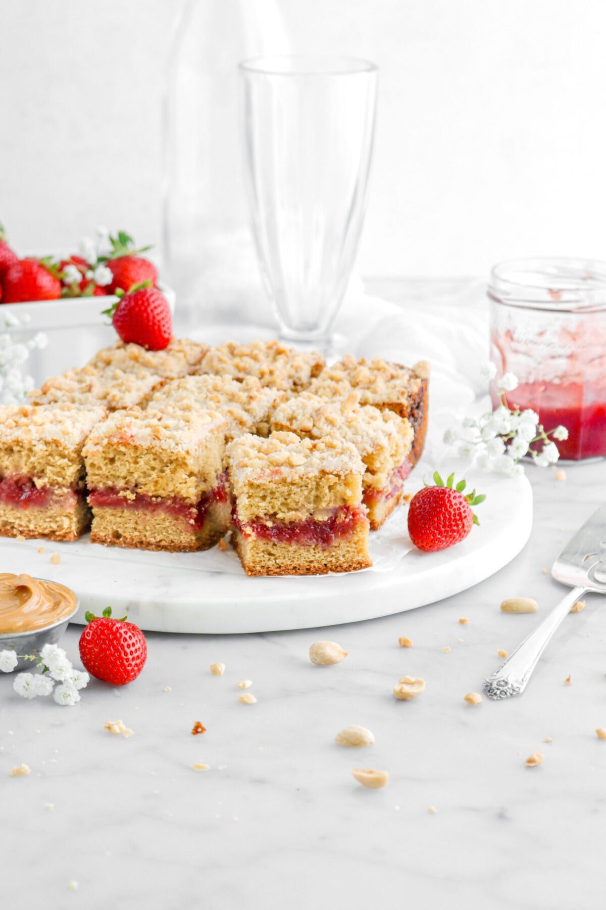 peanut butter and jelly coffee cake square slices on marble surface with white flowers, strawberries, jar of strawberry jam, small bowl of peanut butter, and peanuts around.