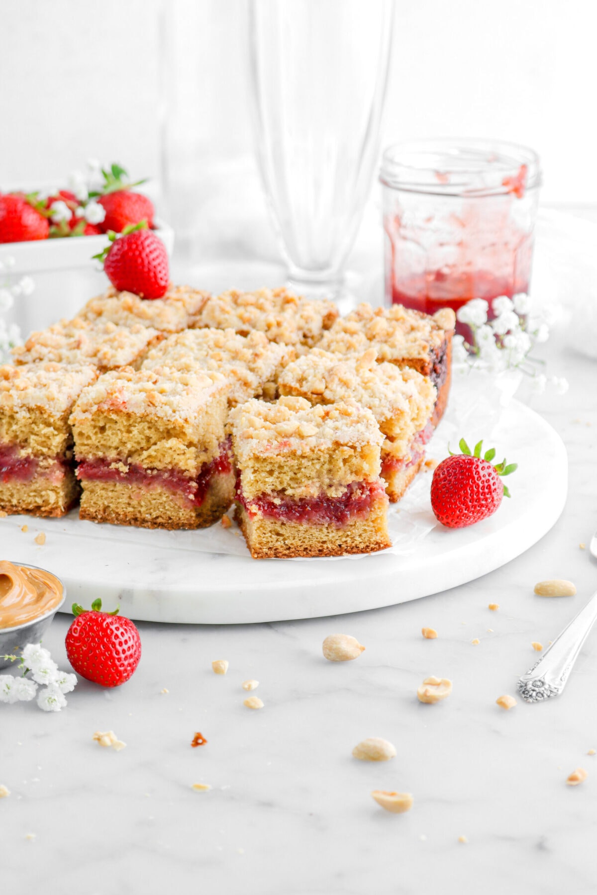 PB&J coffee cake on marble serving tray with fresh strawberries, white flowers, and peanuts around with jar of jam and empty glasses behind.