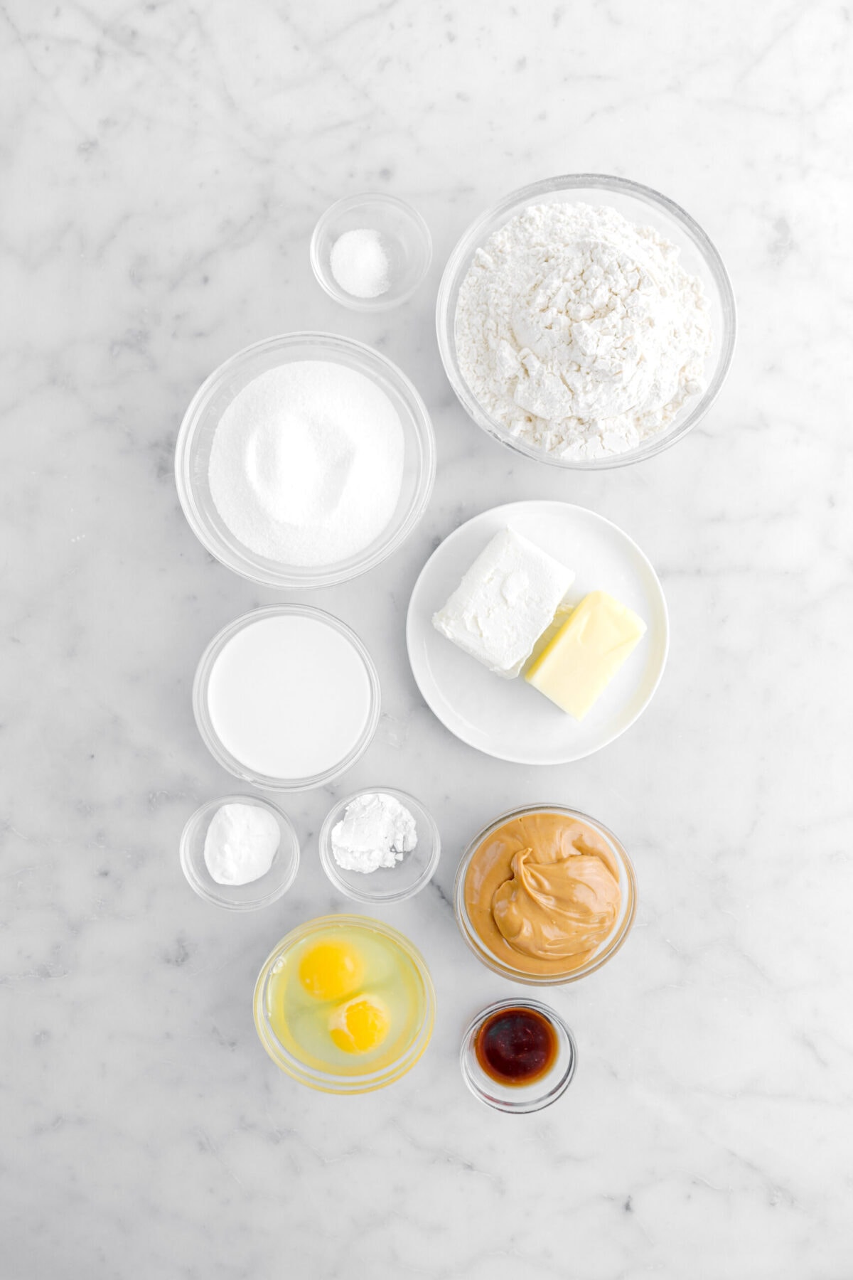 salt, flour, sugar, cream cheese, butter, milk, baking soda, baking powder, peanut butter, eggs, and vanilla on marble surface.