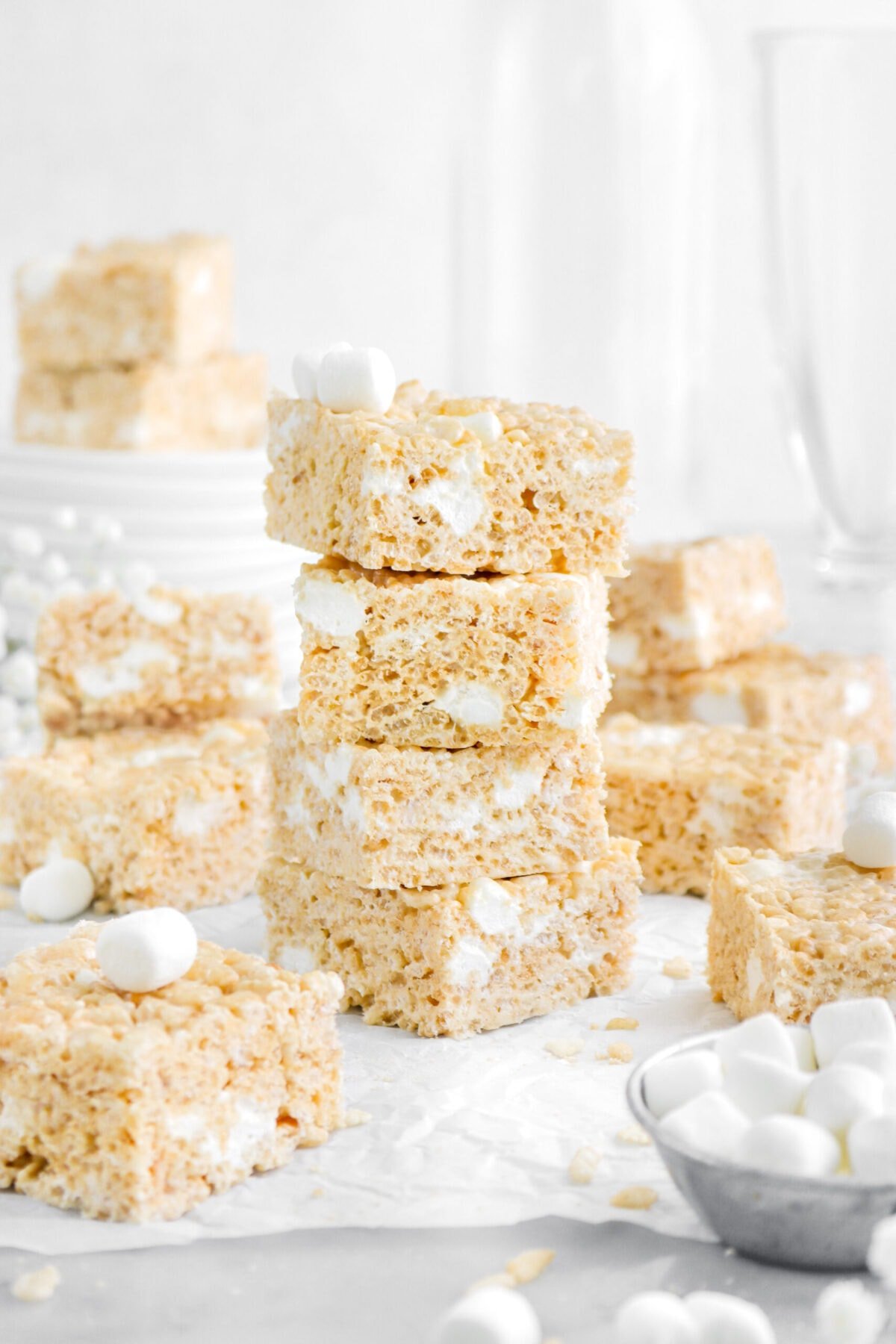 stacked rice krispie treats on parchment paper with more around, a stack of plates behind and two more stacked on top, with glasses behind.