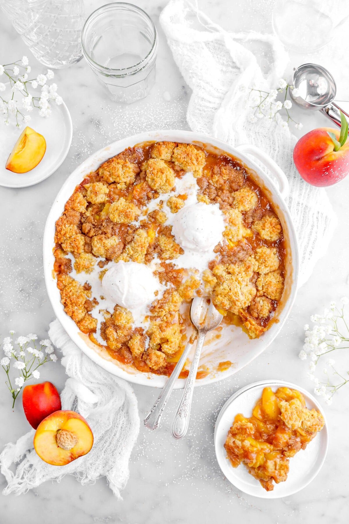 overhead shot of two spoons dug into peach cobbler with two scoops of vanilla ice cream melting atop cobbler with a serving of cobbler on small white plate beside, peaches and flowers around on marble surface.