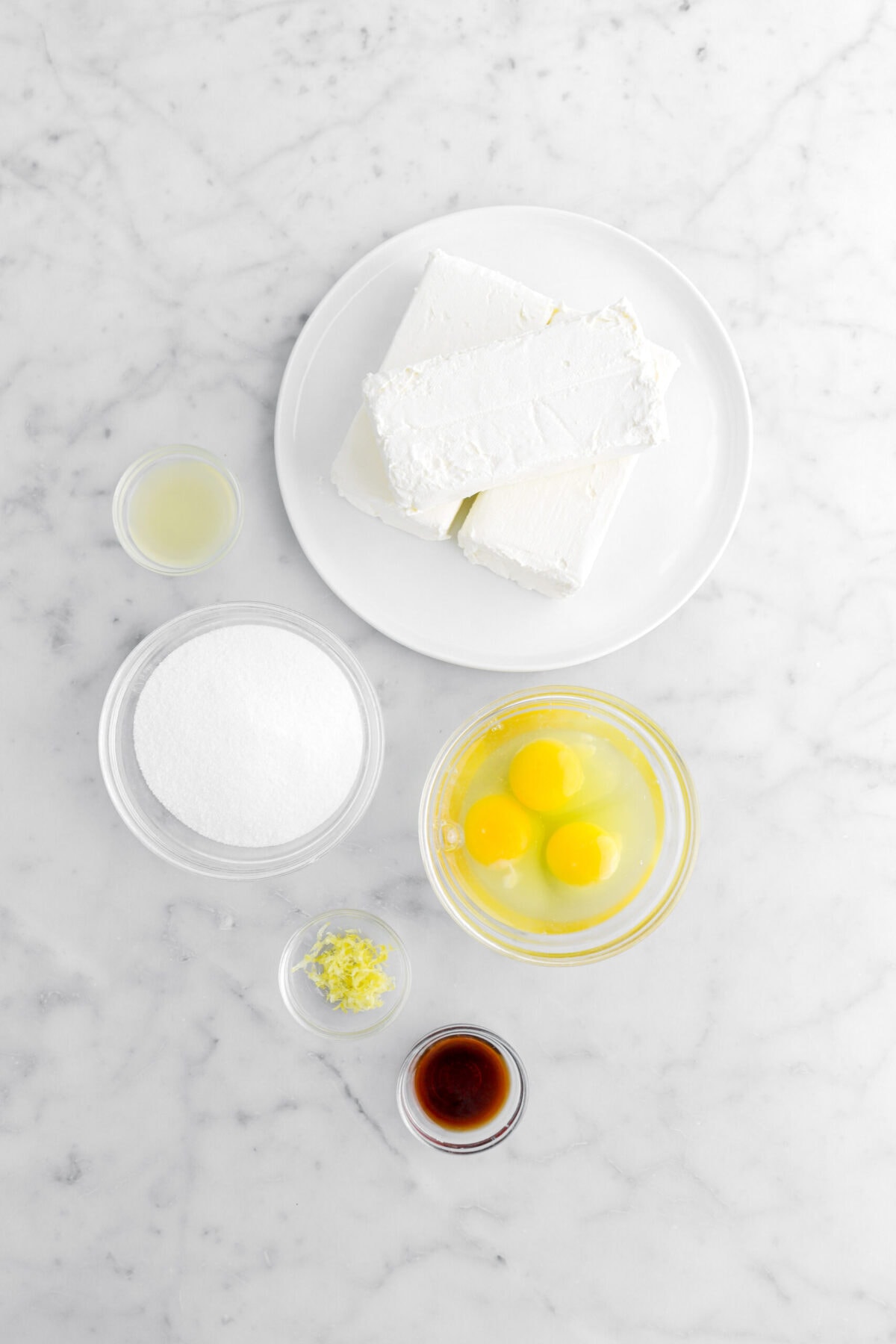 blocks of cream cheese, lemon juice, sugar, eggs, lemon zest, and vanilla on marble surface.
