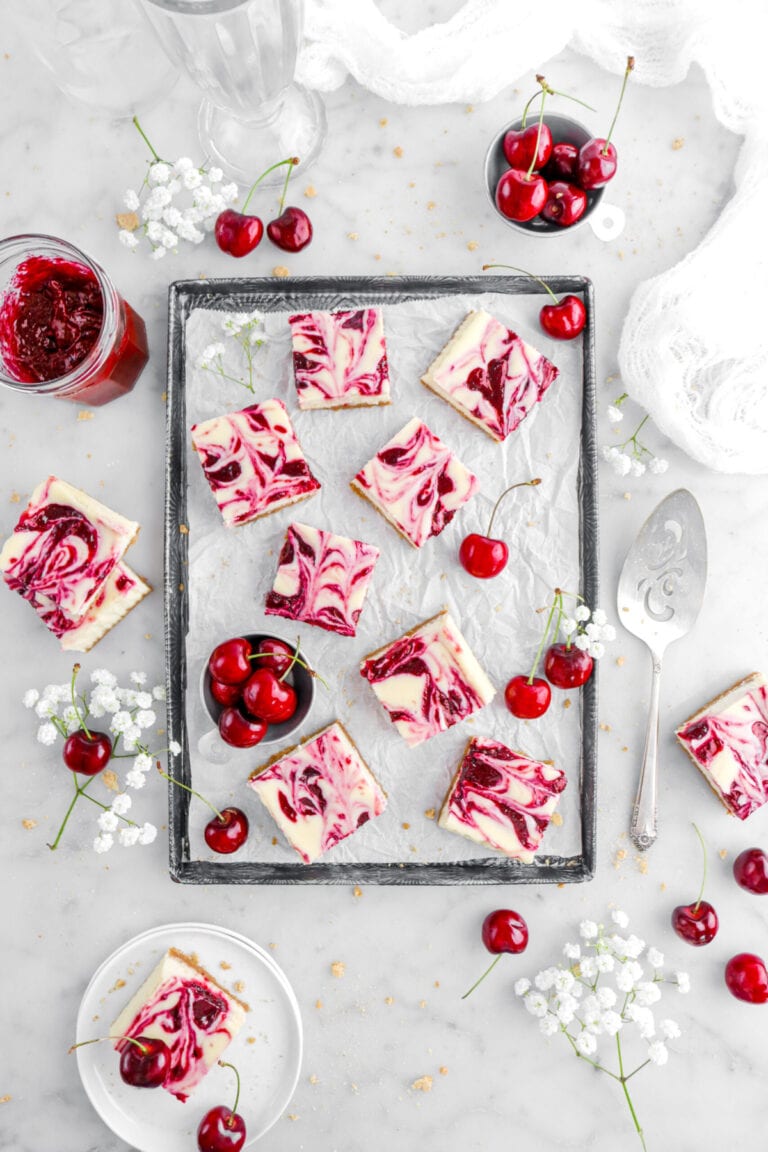 eight cherry swirled cheesecake bars on lined sheet pan with fresh cherries and white flowers around.
