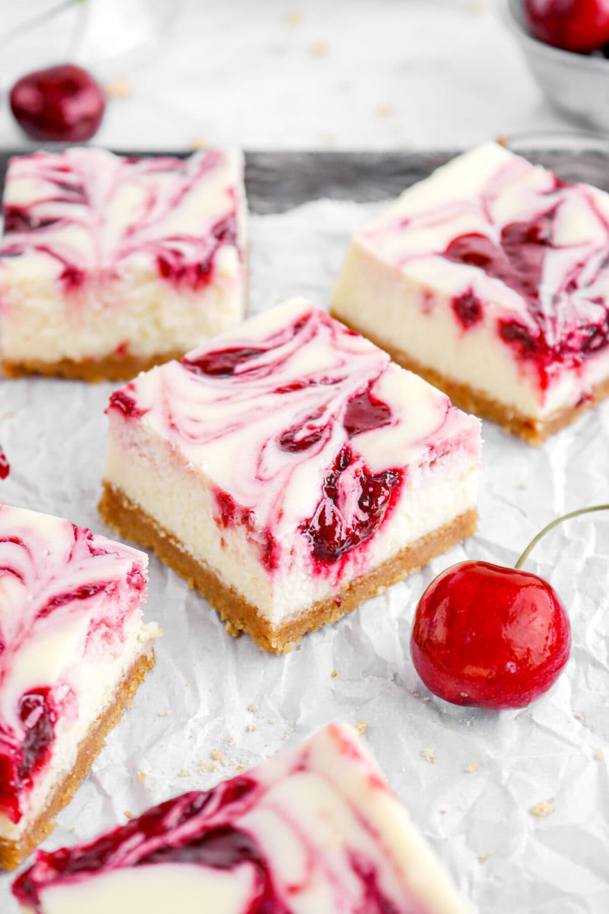 angled close up of cherry swirled cheesecake bar with more cheesecake bars around and a cherry beside.