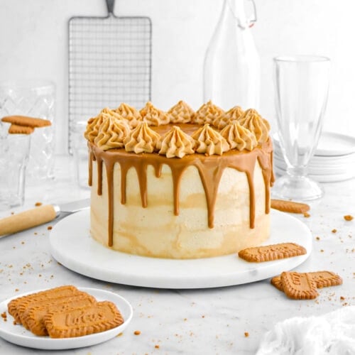 cookie butter cake on marble plate with cookie butter drips and frosting piped around the edges on top of the cake with biscoff cookies around cake, a white cheesecloth, empty glasses, a stack of plates, and a cooking rack behind.