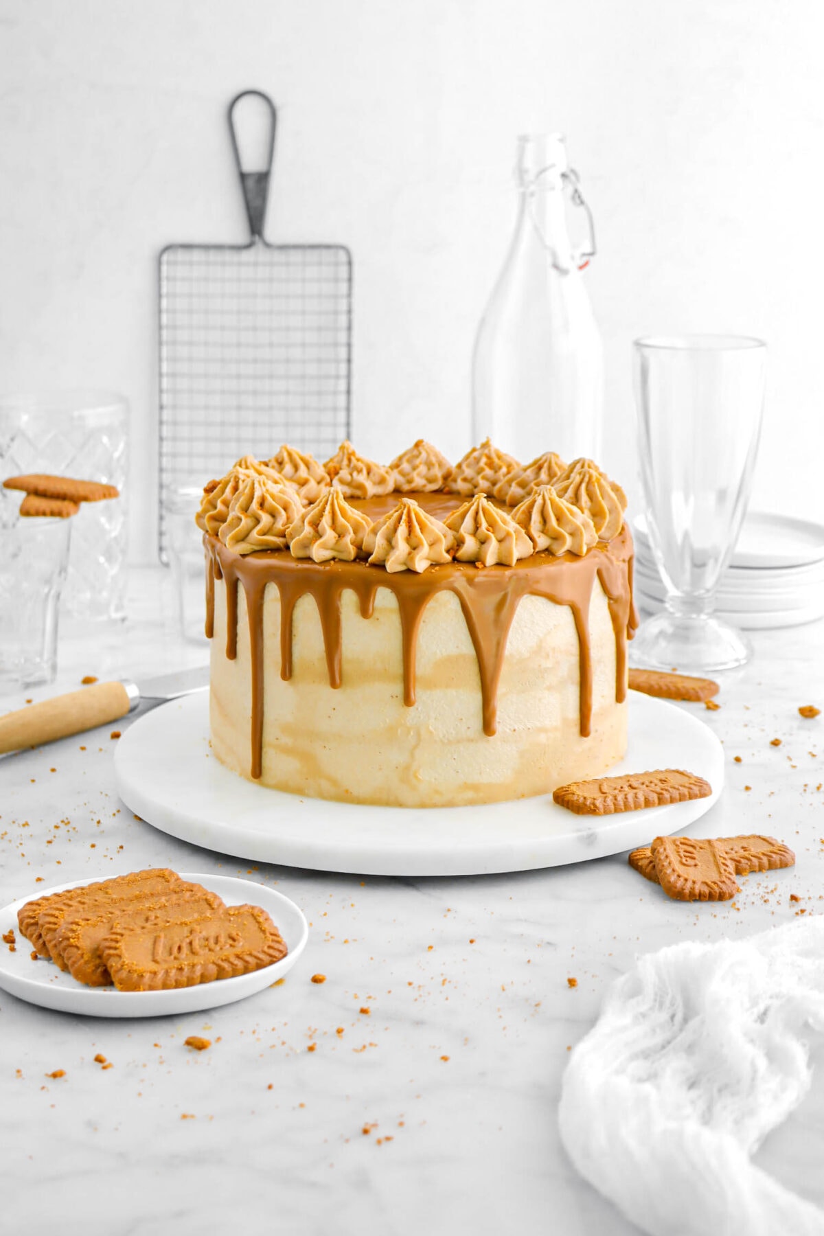 cookie butter cake on marble plate with cookie butter drips and frosting piped around the edges on top of the cake with biscoff cookies around cake, a white cheesecloth, empty glasses, a stack of plates, and a cooking rack behind.