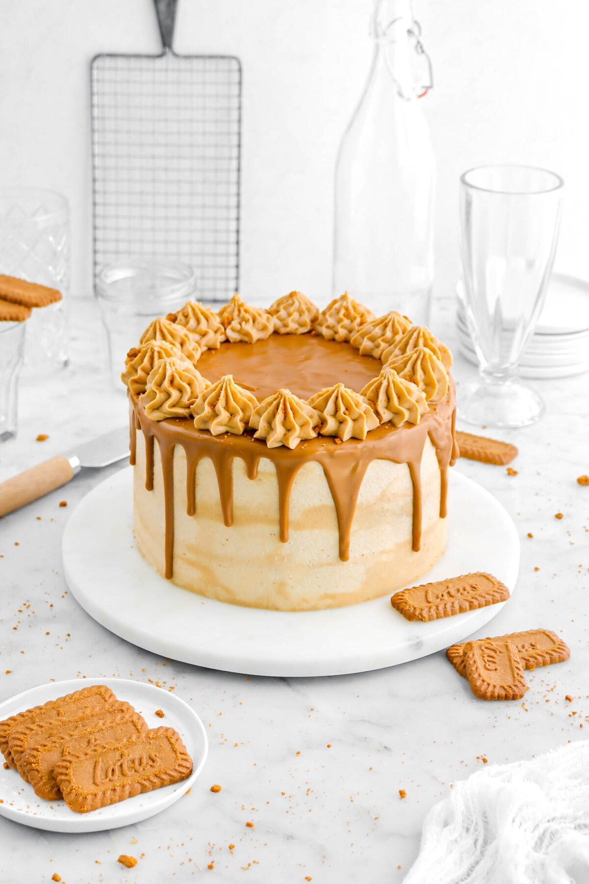 angled shot of cookie butter cake on marble plate with biscoff cookies around, a wood handled spatula, and glasses behind.