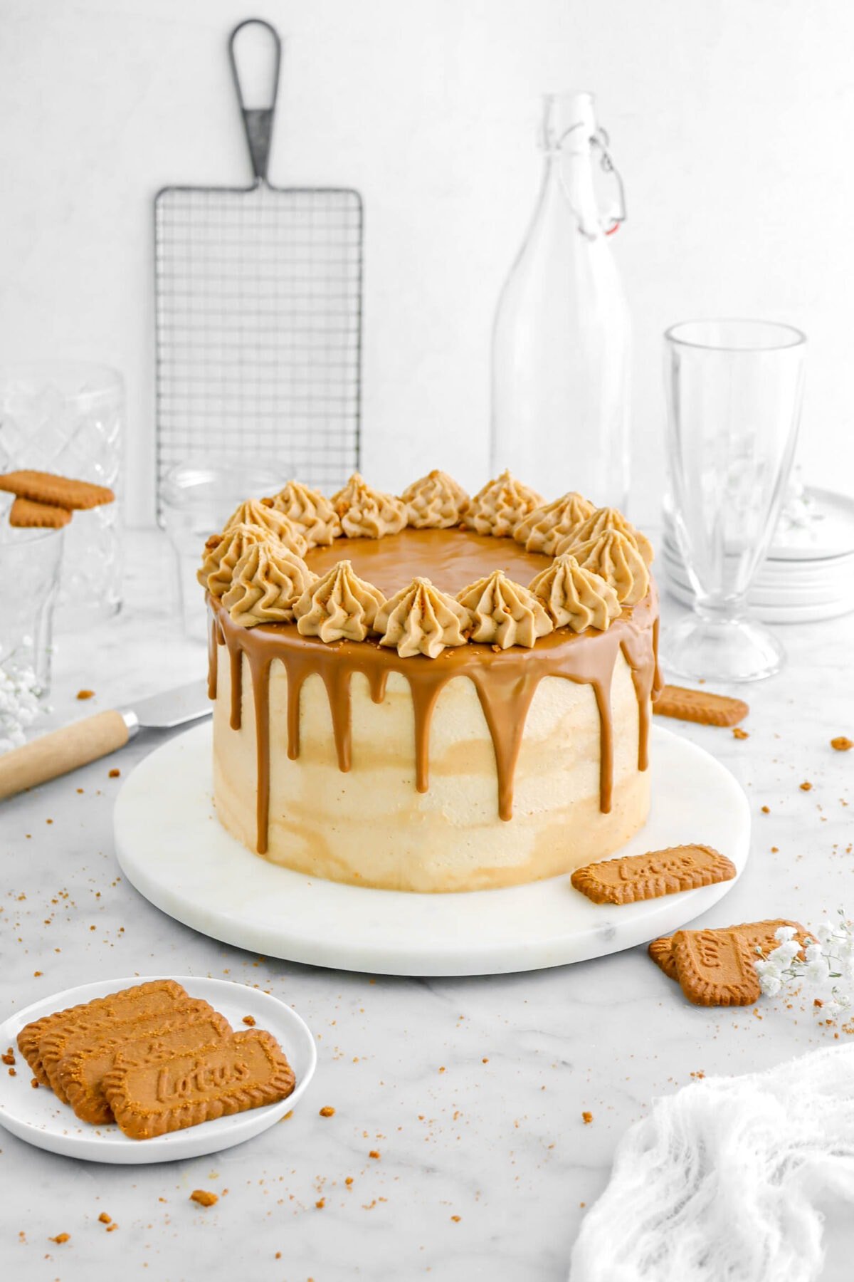 cookie butter cake with cookie butter drips and frosting piped on topm with cookies and cookie crumbs around, empty glasses, and wire tray behind cake on marble surface.