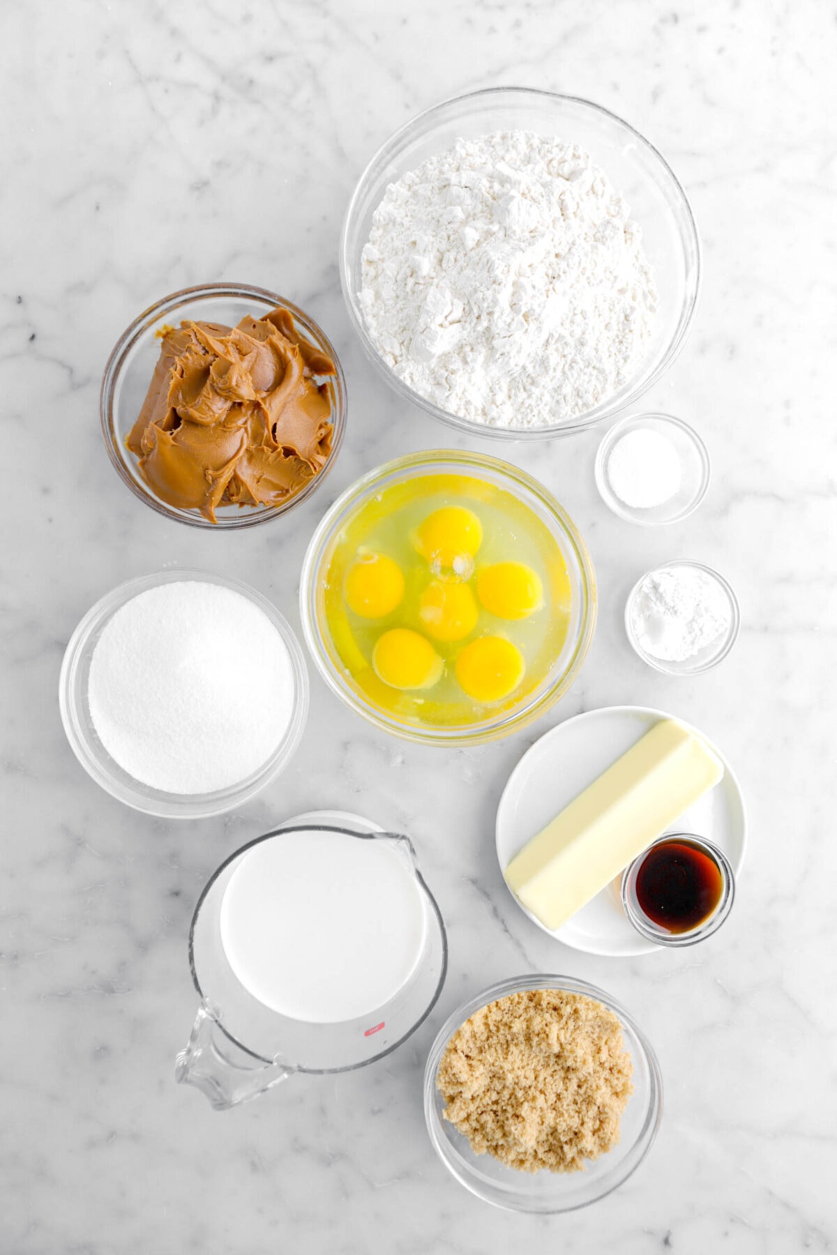 flour, cookie butter, eggs, salt, baking soda, butter, sugar, milk, vanilla, and brown sugar on marble surface.