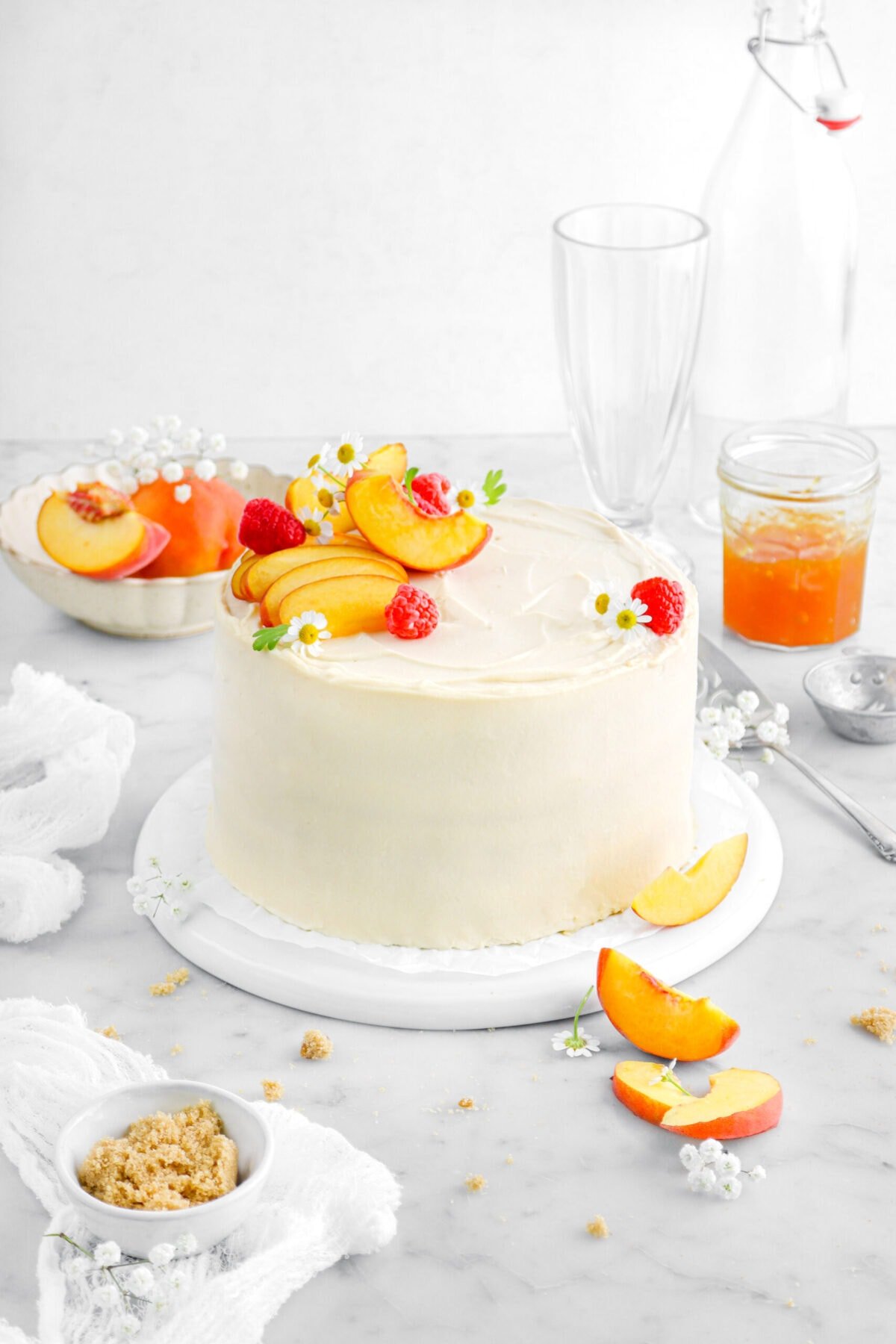 peach cake on upside down white plate with peach slices, raspberries, and chamomile flowers on top of cake, with more peach slices around, a white cheesecloth beside, a jar of peach jam, and two empty glasses behind.