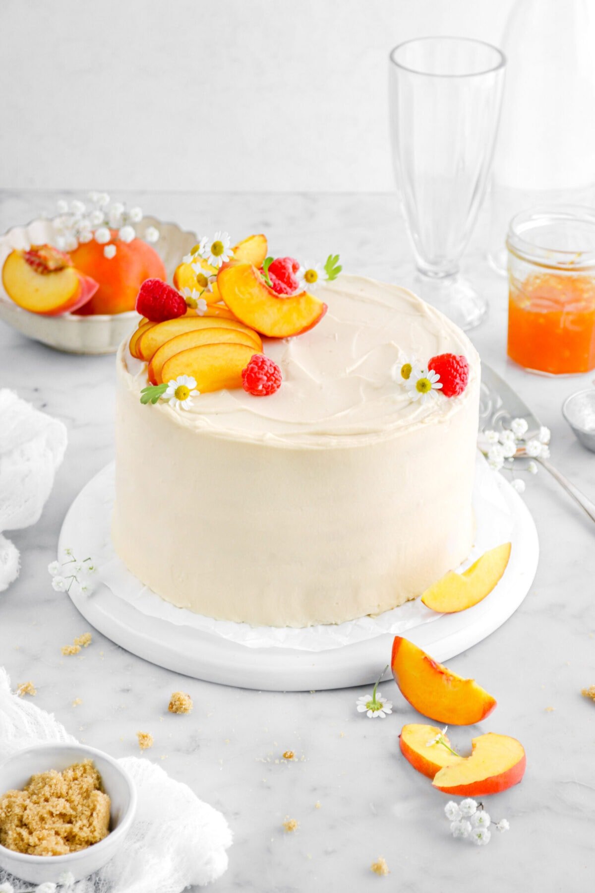 angled close up of peach cake with peach slices, raspberries, and white chamomile flowers on top, with brown sugar and more peach slices around on marble surface.