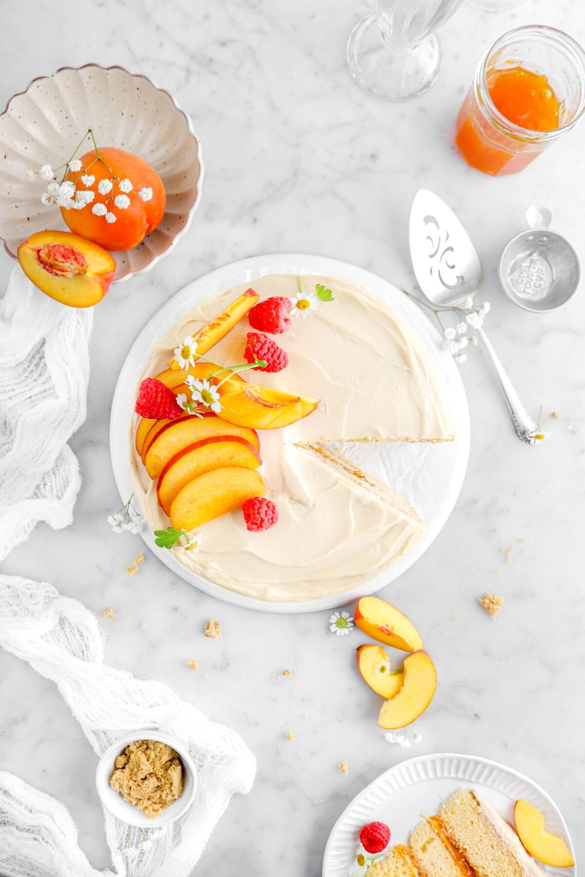 overhead shot of peach cake on white plate with slice of cake on another plate in front, with peaches and flowers around, a cake knife, and peach jam in jar beside on marble surface.