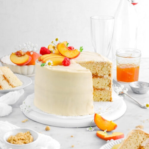 peach cake with two slices in front and behind on white plates with peaches and flowers around on marble surface, a jar of peach jam, and two empty glasses behind cake.