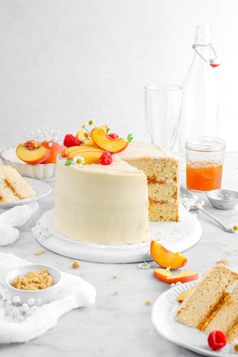 peach cake with two slices in front and behind on white plates with peaches and flowers around on marble surface, a jar of peach jam, and two empty glasses behind cake.