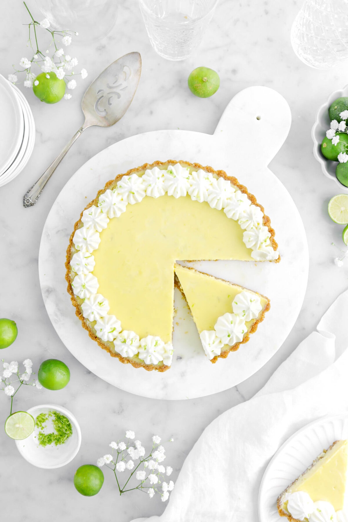 overhead shot of key lime pie with slice cut into it on marble surface with key limes and white flowers around.