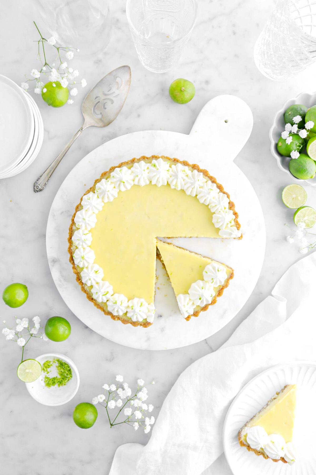 key lime pie with slice cut into it with another slice on white plate beside with key limes and white flowers around on marble surface.