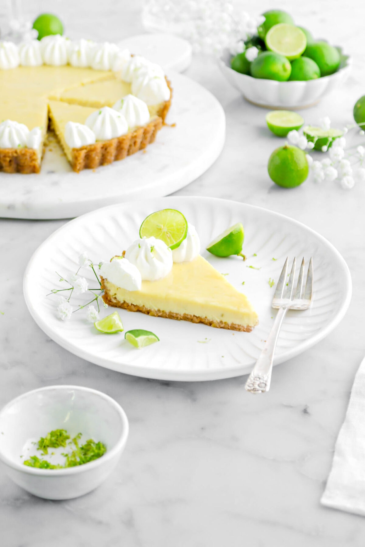 slice of key lime pie on white plate with fork beside, key lime pieces, and white flowers around, with pie and key limes behind on marble surface.