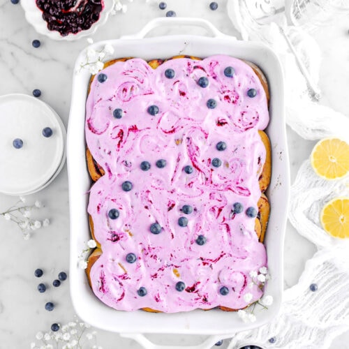 frosted blueberry rolls in white casserole with blueberries and two lemon halves around on marble surface.