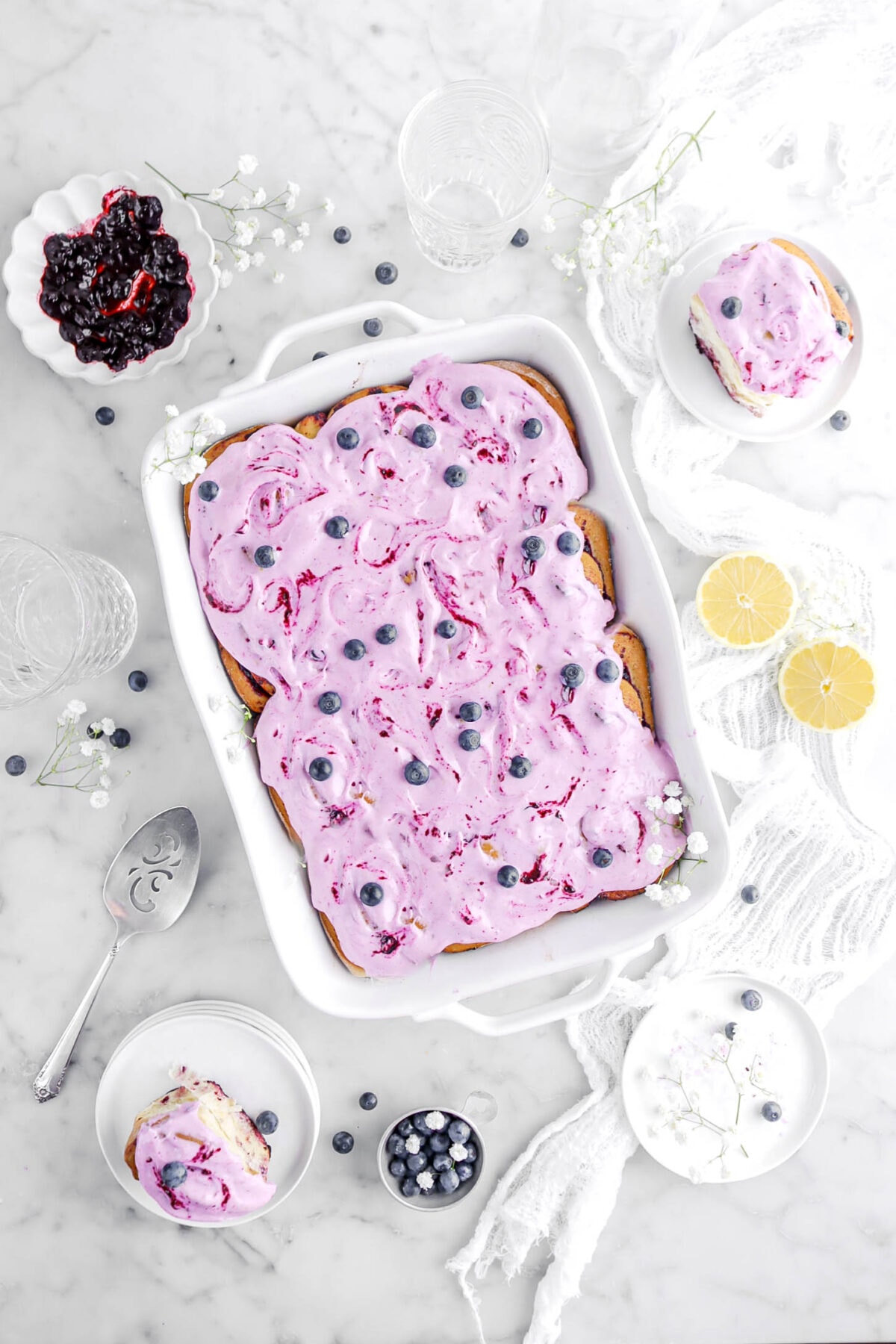 overhead shot of lemon blueberry rolls in white casserole with two extra rolls on white plates beside casserole with blueberries, two lemon halves, and white flowers around.