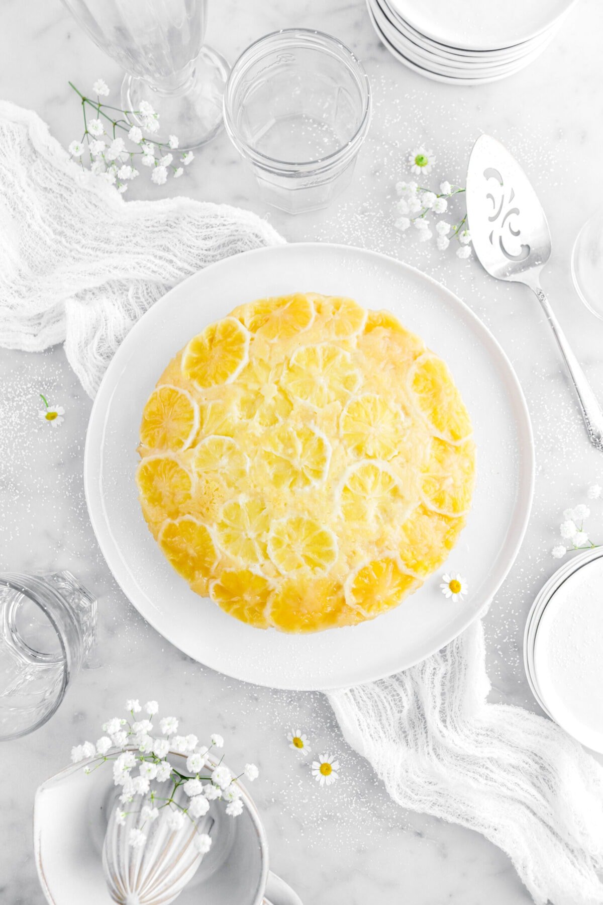 lemon upside down cake on white plate on top of a white cheesecloth with chamomile and other white flowers around on marble surface.