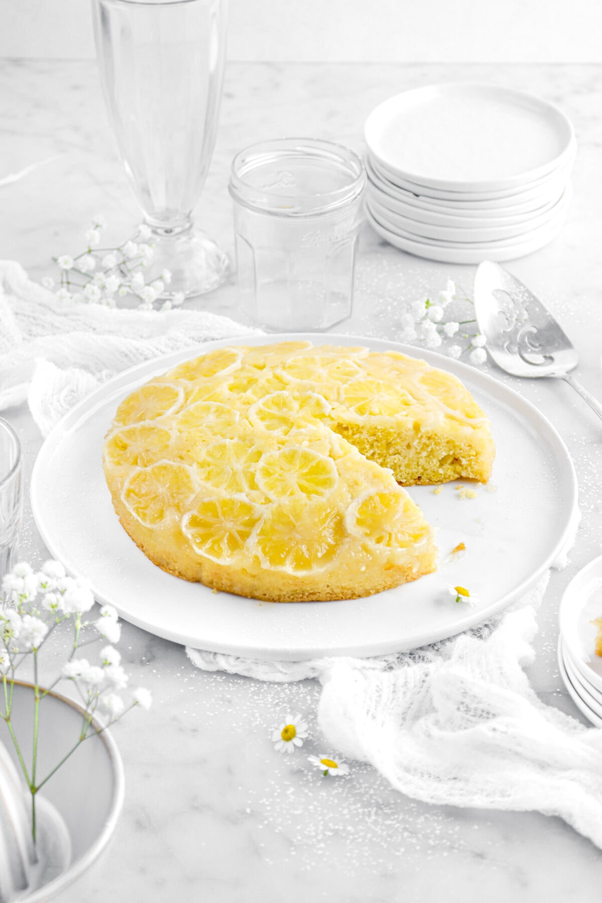 front shot of lemon upside down cake with slice missing with stack of plates and empty glasses behind with flowers around on marble surface.