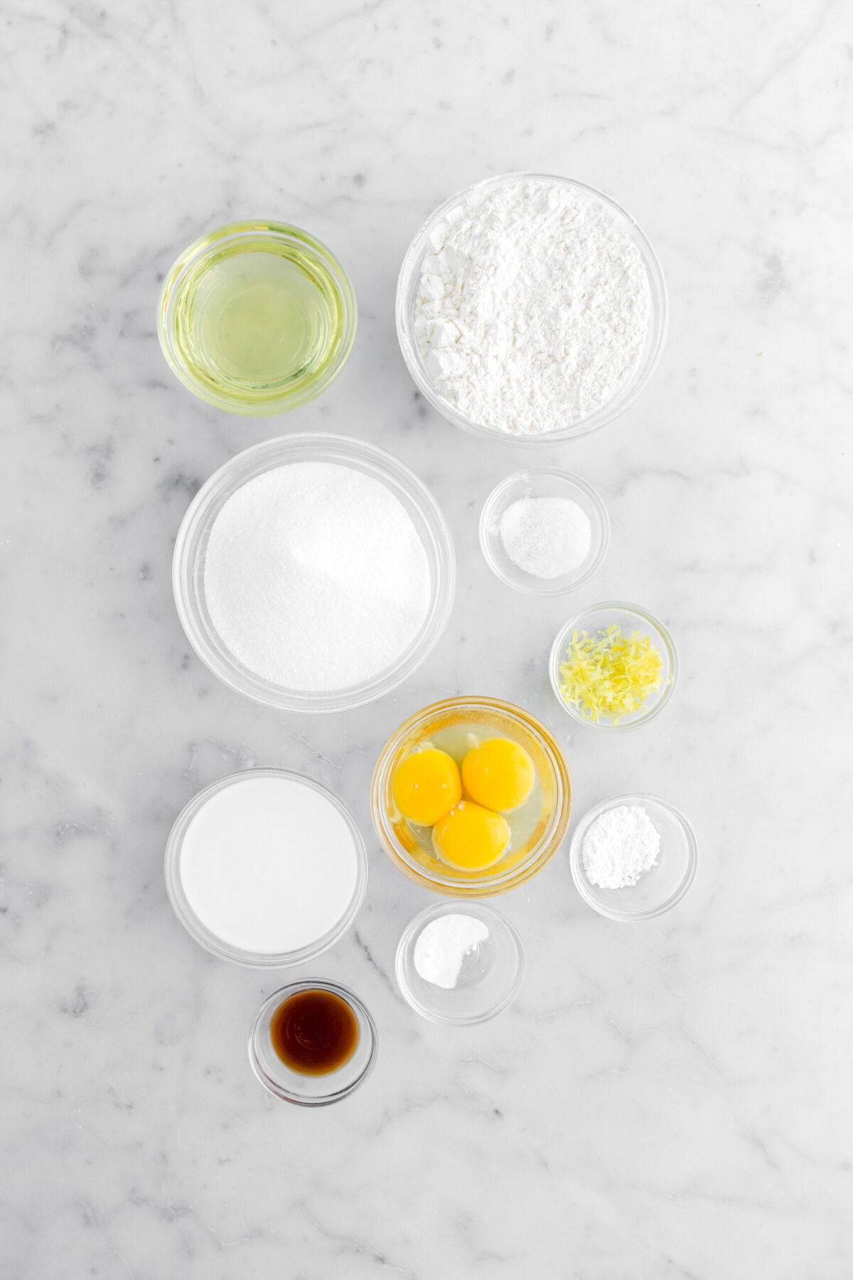 oil, flour, sugar, salt. lemon zest, eggs, baking powder, baking soda, milk, and vanilla on marble surface.