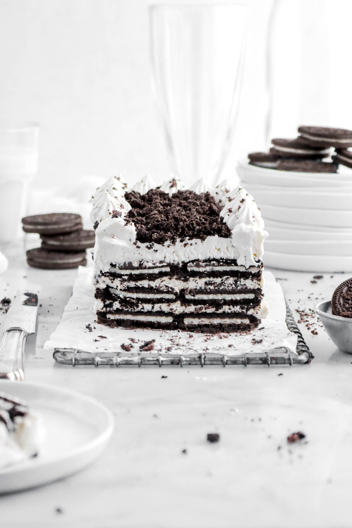 cut oreo ice box cake with white plate in front with slice on top on marble surface with oreo crumbs and oreos around.
