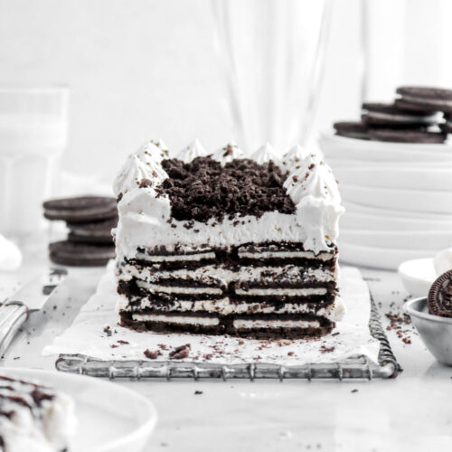 oreo icebox cake on wire tray with slice of cake on small white plate beside, with oreos around, a glass of milk behind, a stack of white plates, and two empty glasses.
