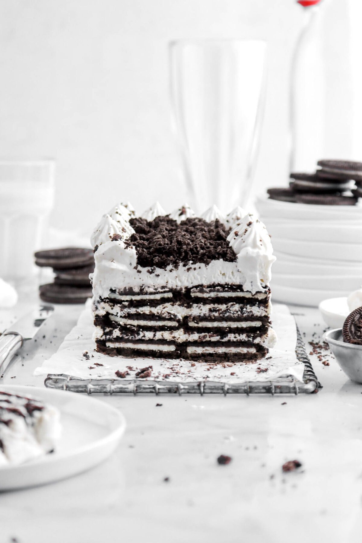 oreo icebox cake on wire tray with slice of cake on small white plate beside, with oreos around, a glass of milk behind, a stack of white plates, and two empty glasses.