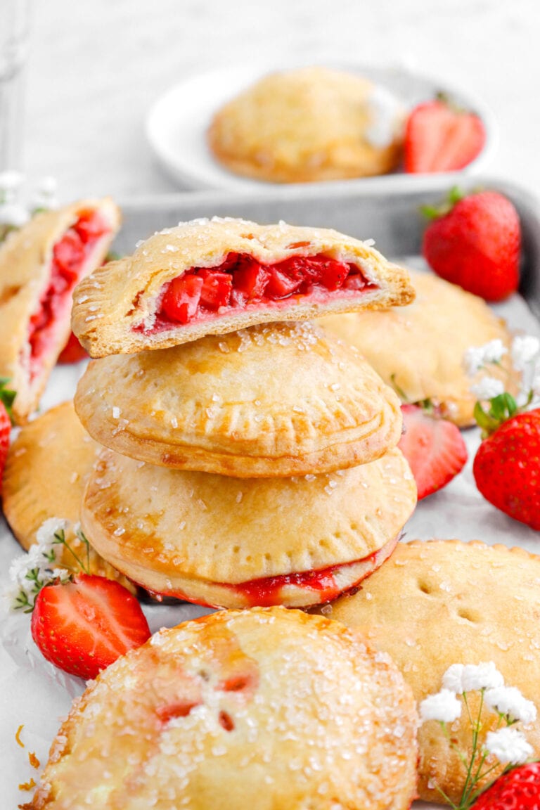 three stacked strawberry hand pies with top hand pie cut in half with more hand pies and strawberries around.