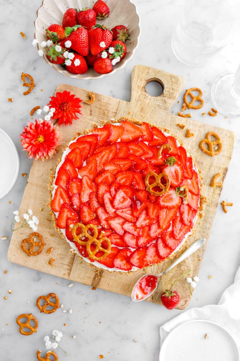 strawberry tart on square wood serving board with pretzels, strawberries, and flowers around on marble surface and spoon of jam beside tart.