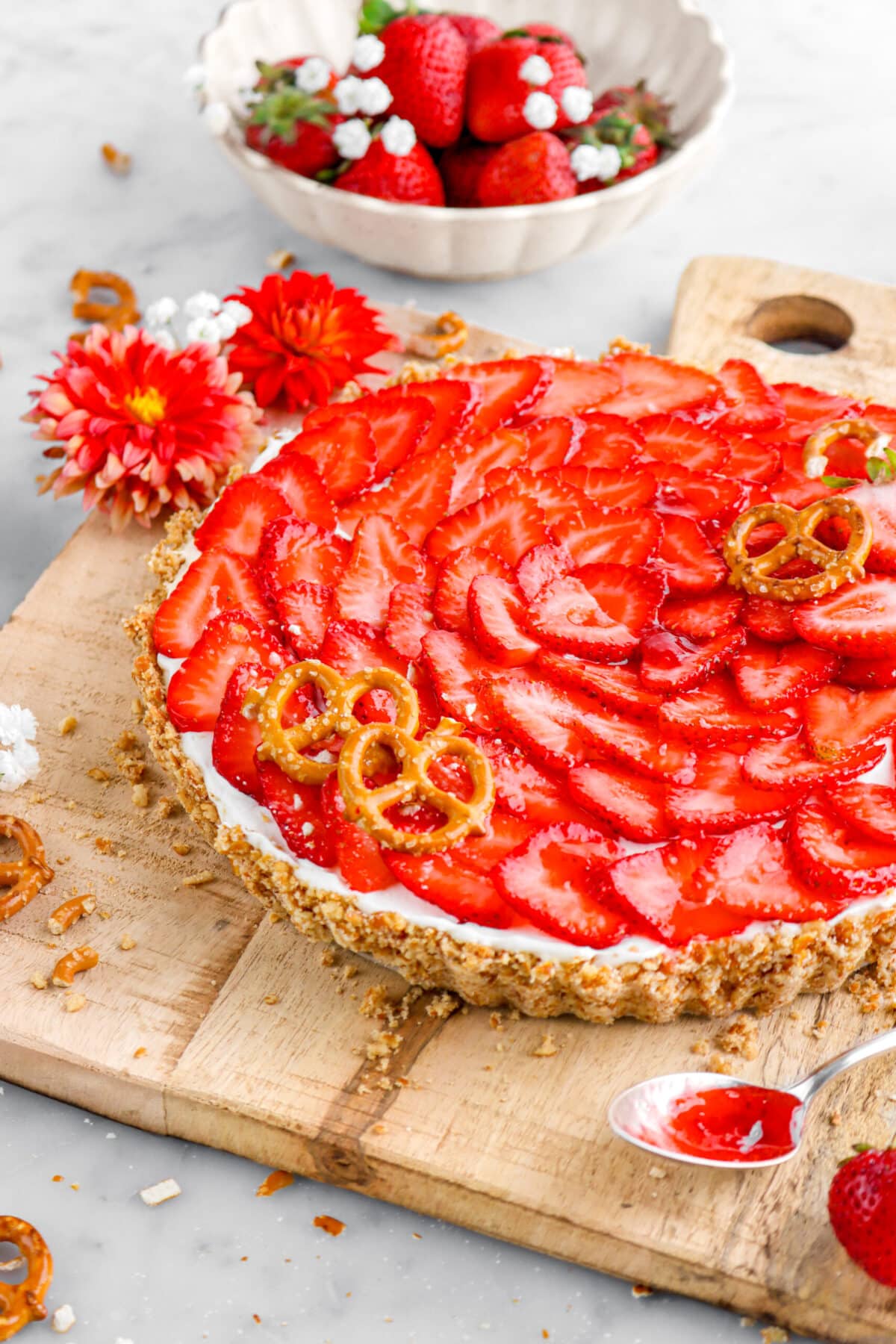 cropped close up of strawberry tart on wood board with flowers around and bowl of strawberries behind.
