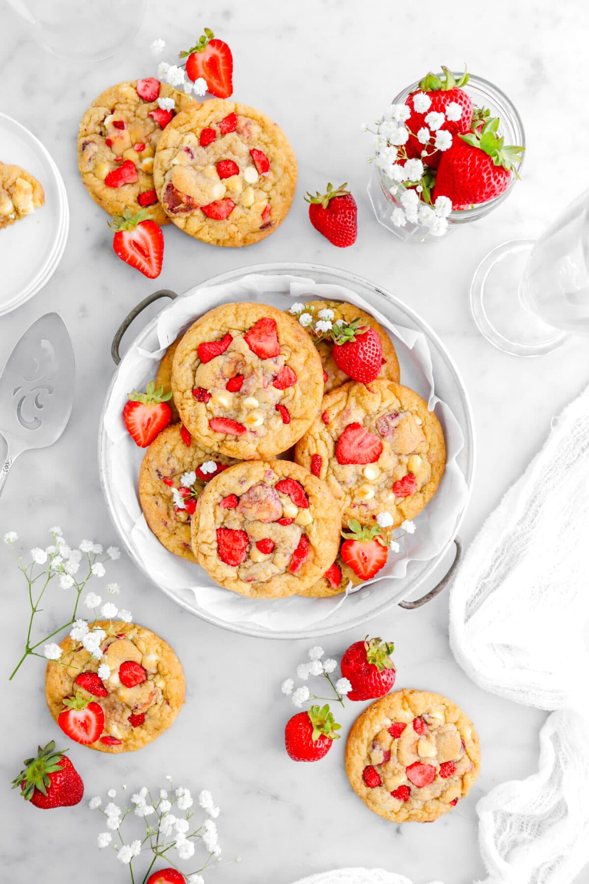 six cookies in pie plate with four more cookies around, white flowers, and strawberries on marble surface with cheesecloth and cake knife beside.