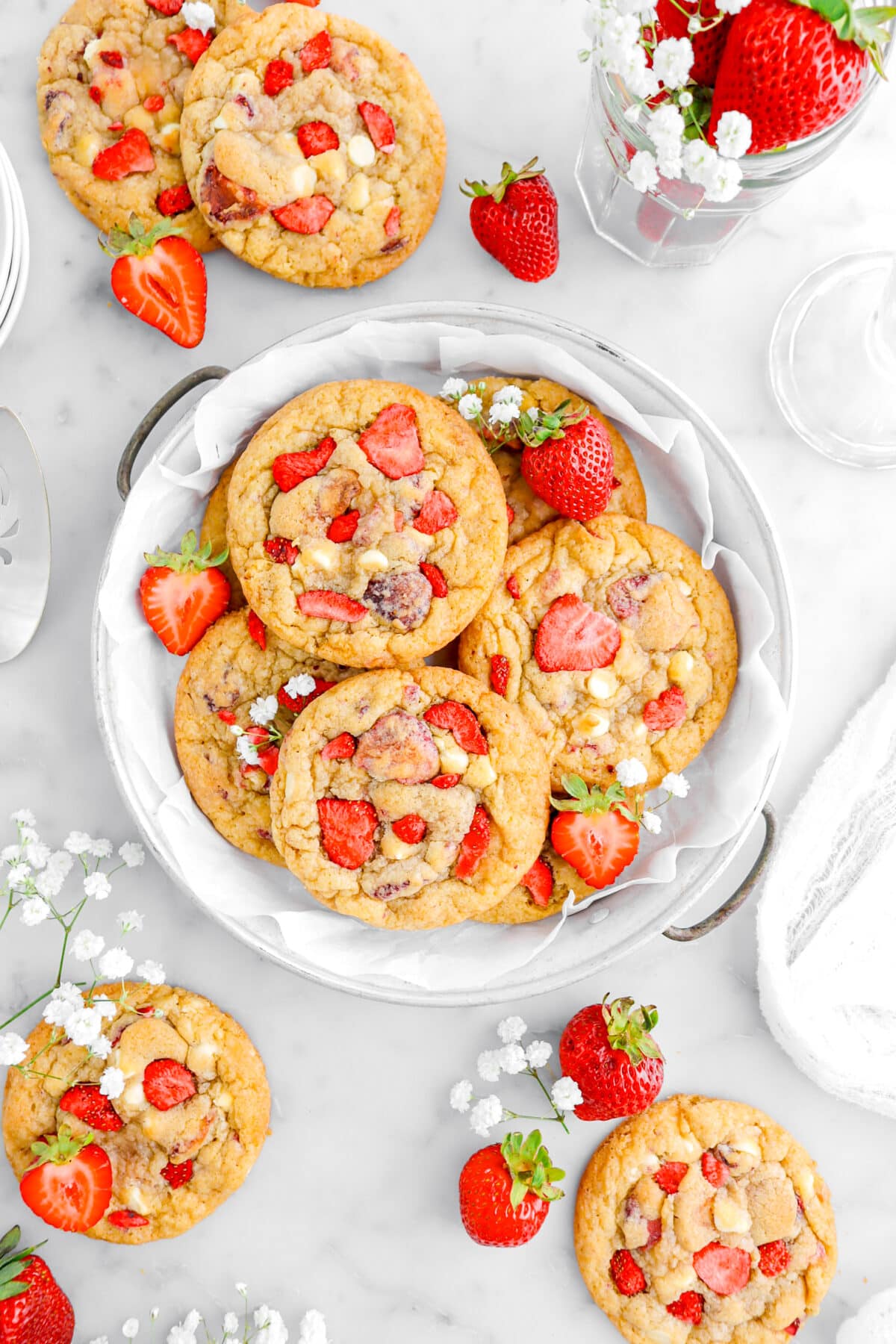 overhead shot of six white chocolate chip and strawberry cookies in lined pie plate with four more cookies around, fresh strawberries, and white flowers on marble surface.