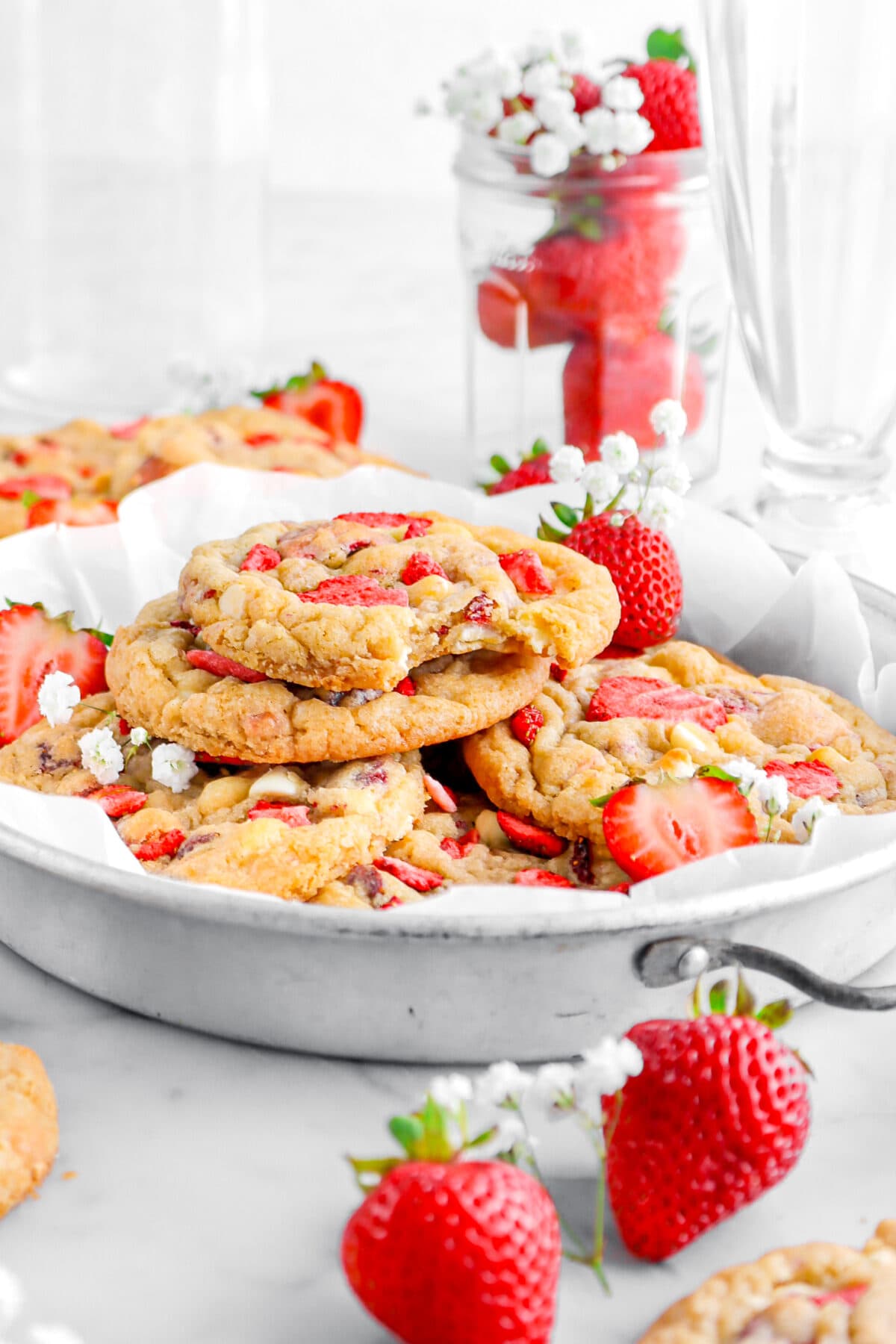 stacked cookies in pie plate with top cookie missing a bite with jar of fresh strawberries and white flowers behind.