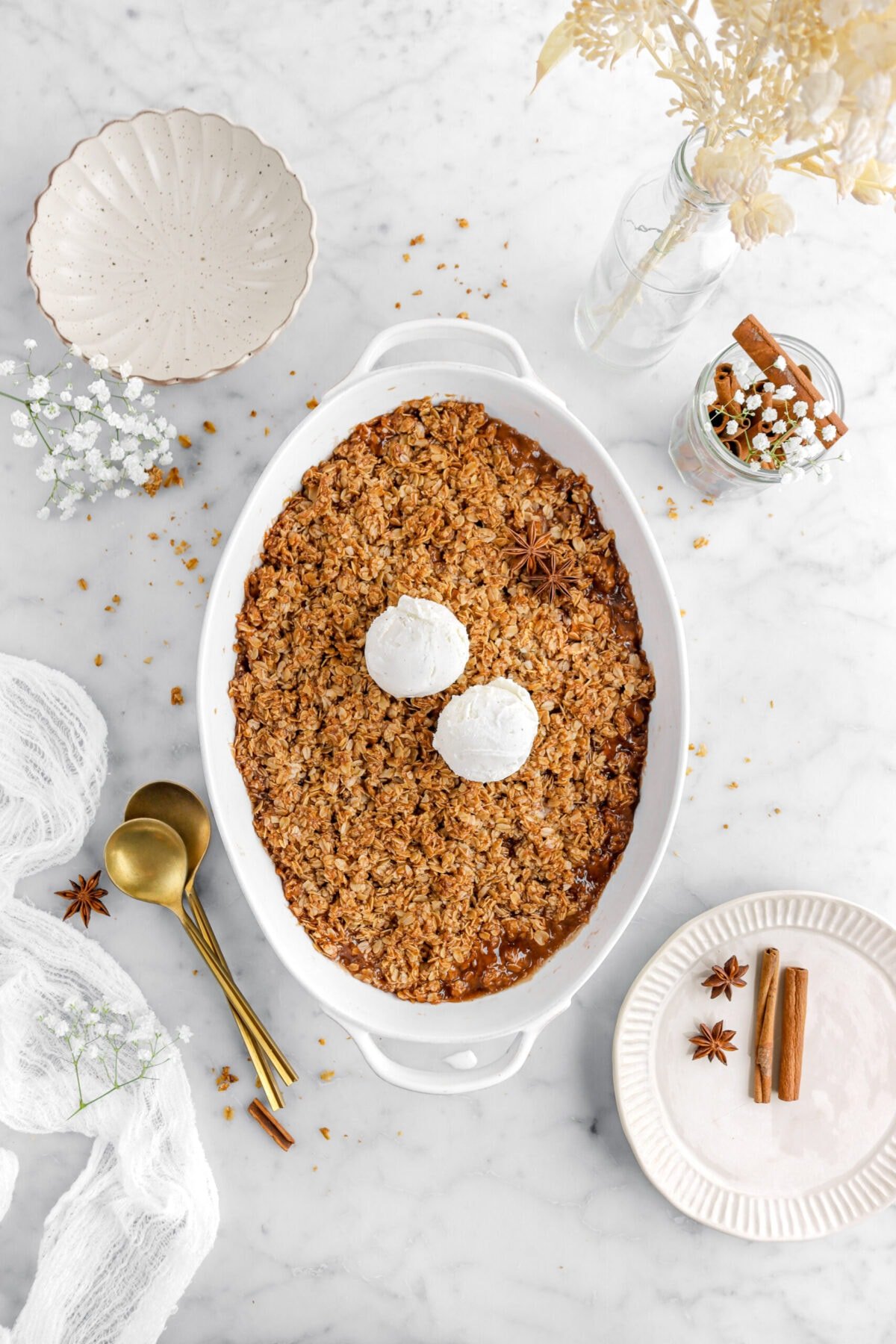 apple crisp with two scoops of ice cream on top with a plate, whole spices, and an empty bowl around on marble surface with two gold spoons beside.
