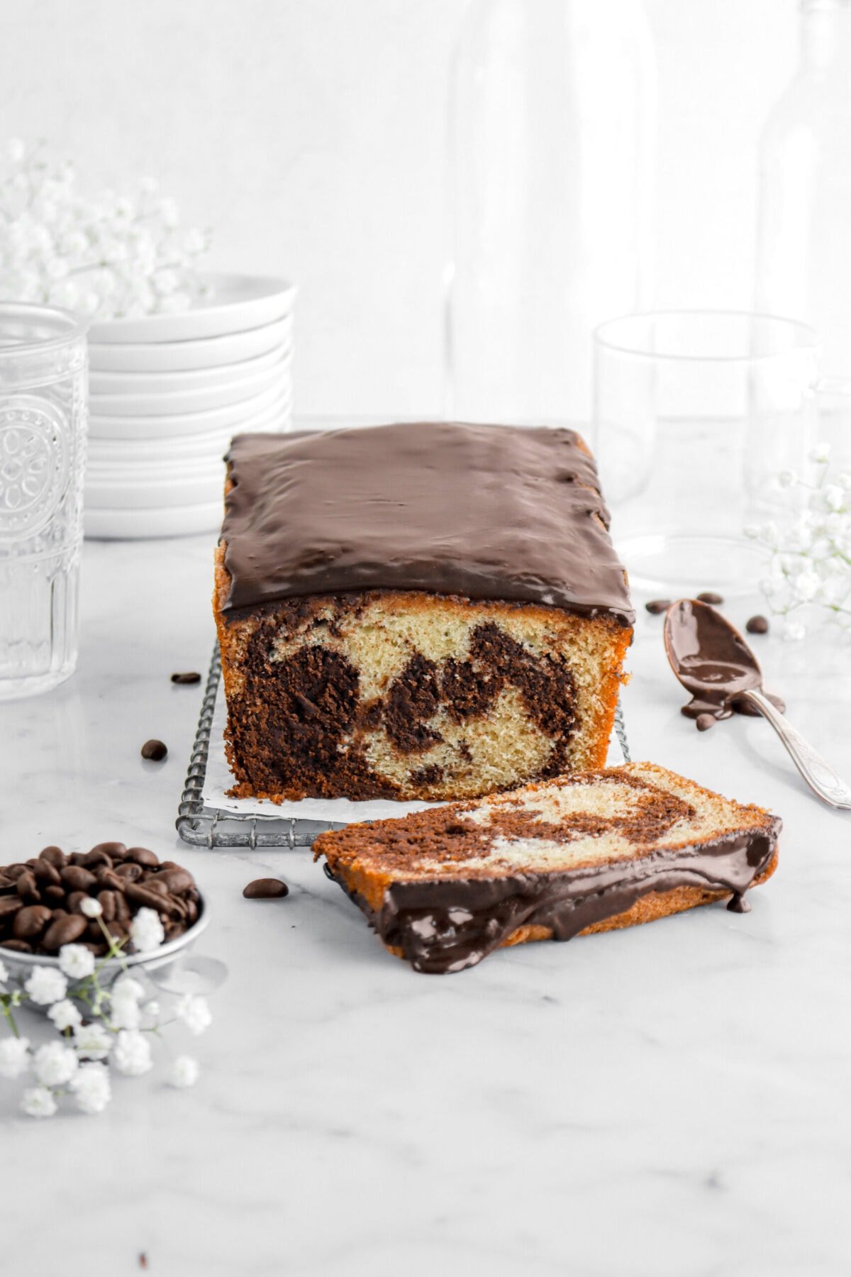 marble loaf cake on small wire tray with slice laying in front, measuring cup of coffee beans and spoonful of chocolate glaze beside with flowers around on marble surface.