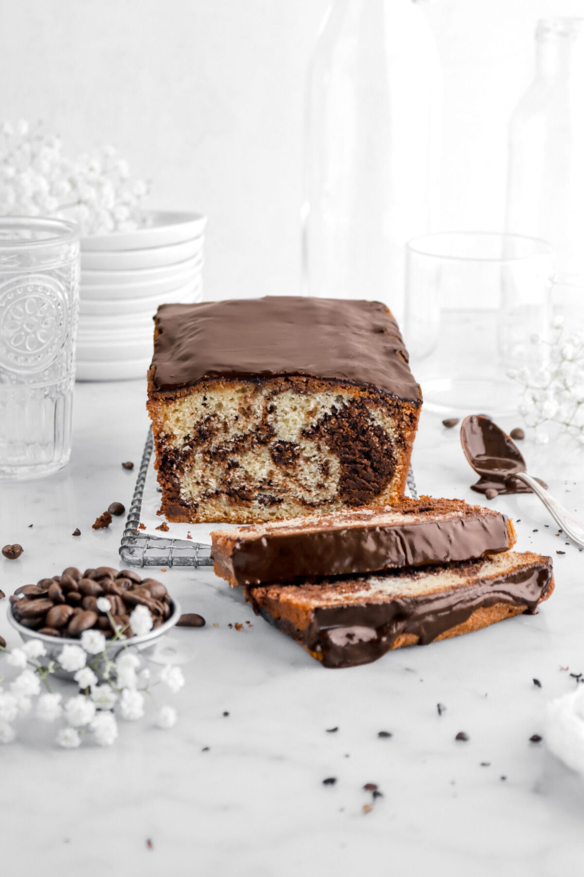 chocolate marble cake on parchment lined wire tray with two slices laying on front with spoon of chocolate icing and bowl of coffee beans beside with more coffee beans scattered around, white flowers, a stack of plates, and empty glasses around.