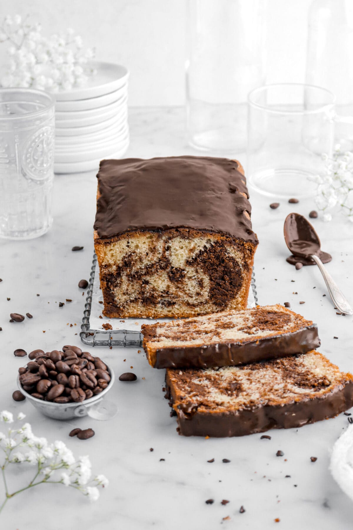 angled shot of chocolate marble loaf cake with two slices laying in front with coffee beans and white flowers around, a spoon of chocolate icing beside, and stack of plates behind.