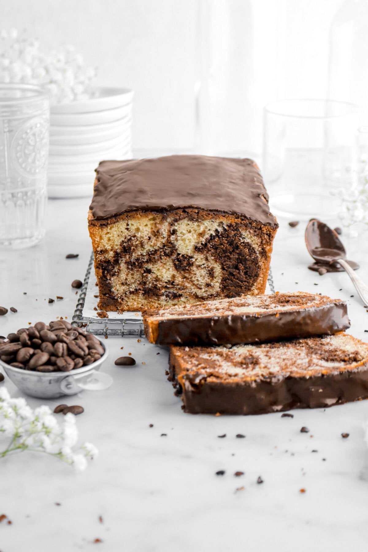 front shot of marble loaf cake with two slices in laying in front with bowl of coffee beans beside, a spoon of chocolate icing, and flowers around.